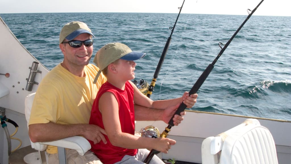 Two people fishing on a boat in the ocean. The adult, wearing sunglasses and a cap, sits behind a child holding a fishing rod. Both appear focused as they enjoy their Costa Rica fishing charter off the picturesque shores of Playa del Coco.